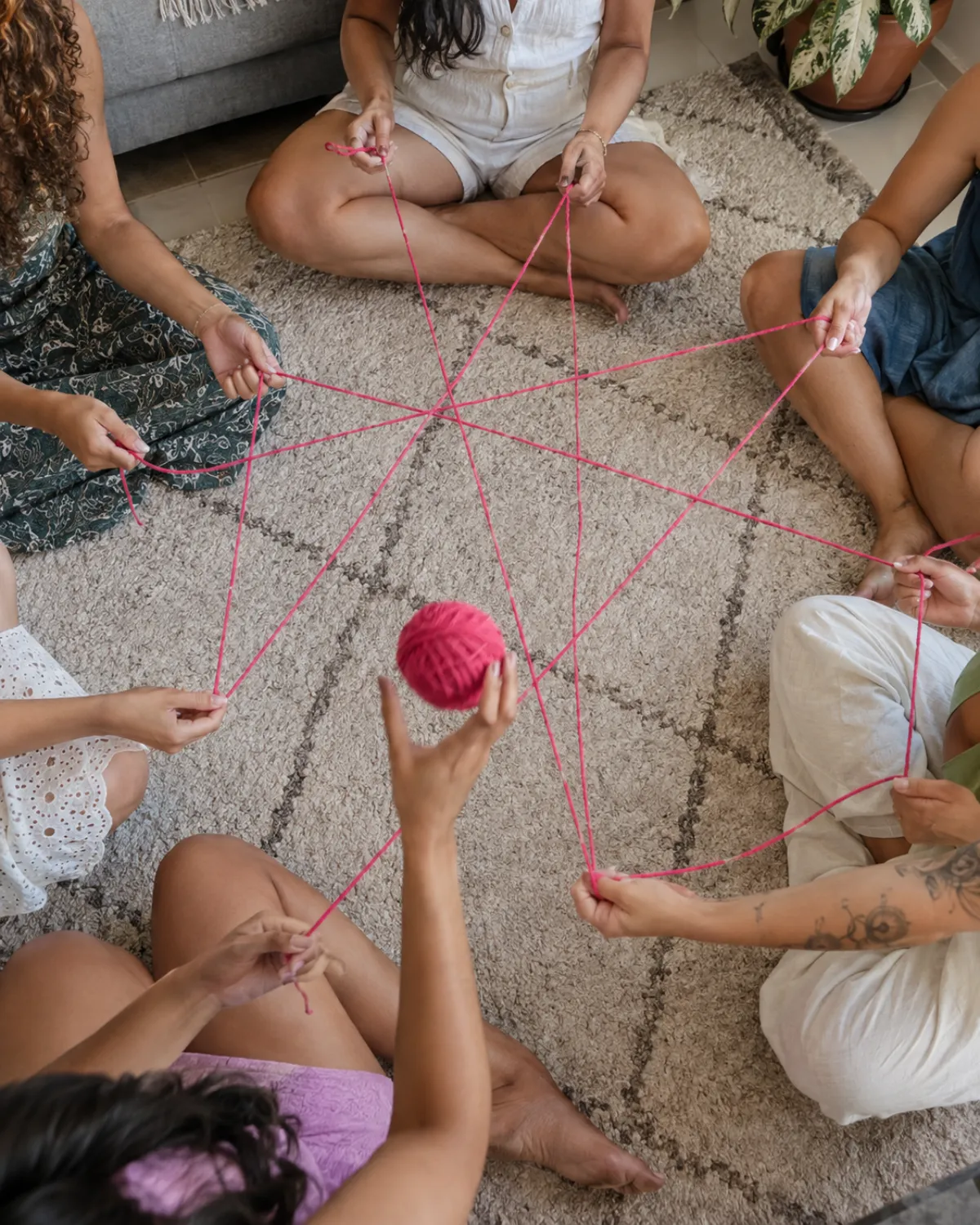 Amigas sentadas em roda no tapete formando uma teia com novelo de lã rosa
