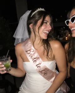 Noiva com tiara de pérolas, véu curto e faixa Bride to Be rindo com amiga de óculos gatinho
