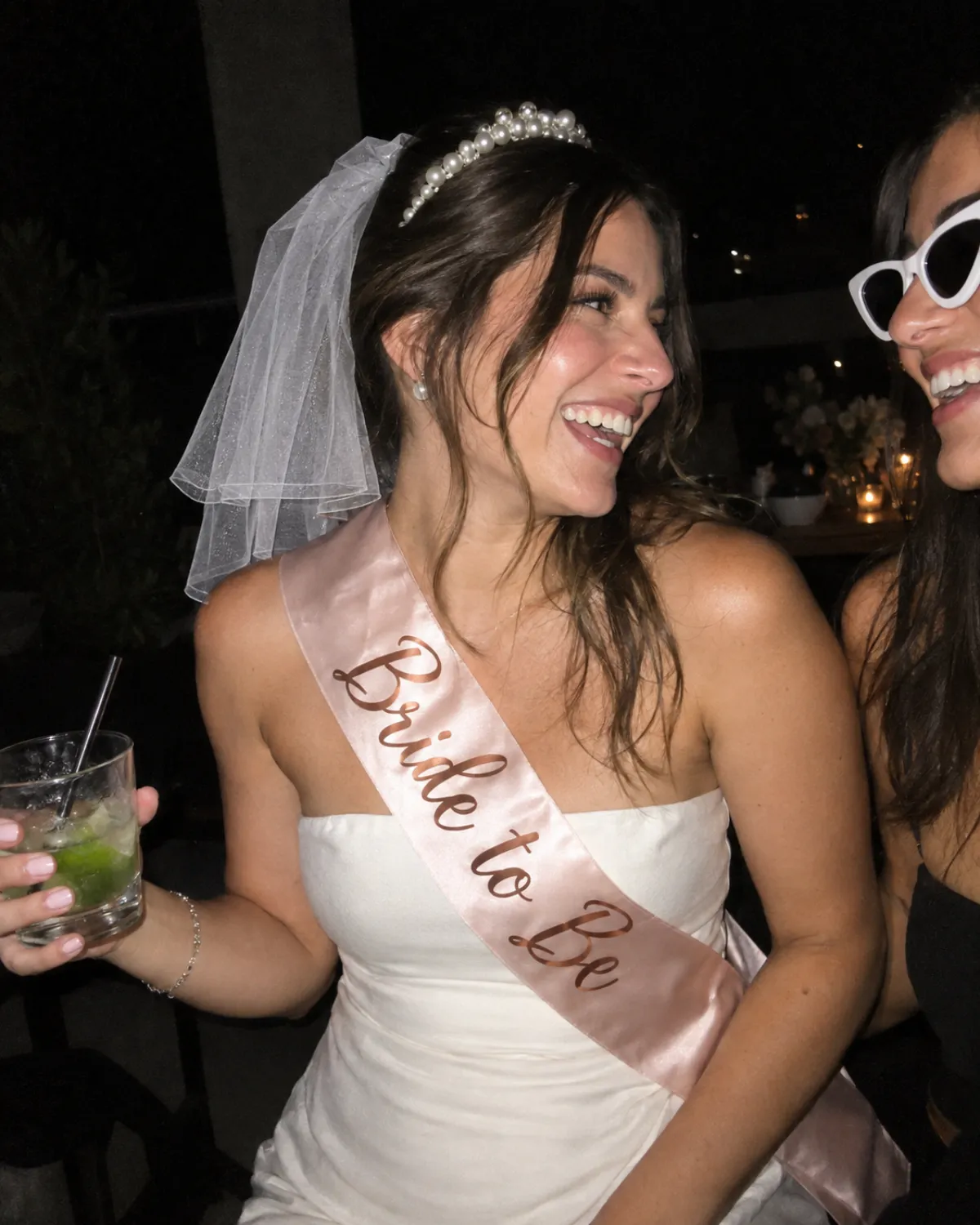 Noiva com tiara de pérolas, véu curto e faixa Bride to Be rindo com amiga de óculos gatinho